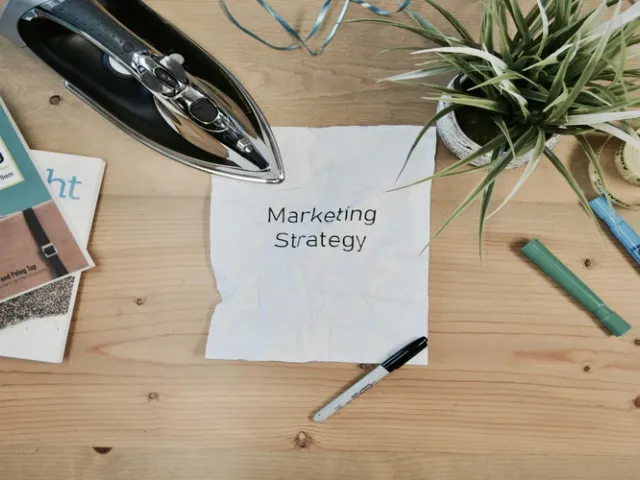 Mesa de escritório com um papel centralizado escrito estratégia de marketing com 3 canetas sobre a mesa, 3 livros que falam sobre marketing, uma fita de medidas e um jarro de planta.