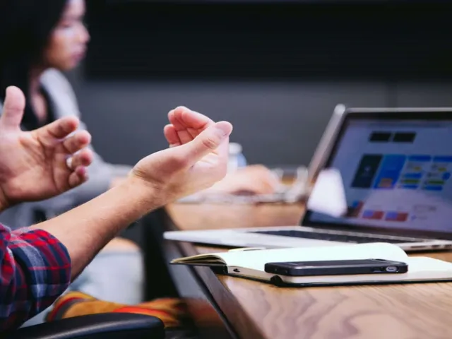 Equipe em escritório com mesa amadeirada com um caderno de anotações e um computador.