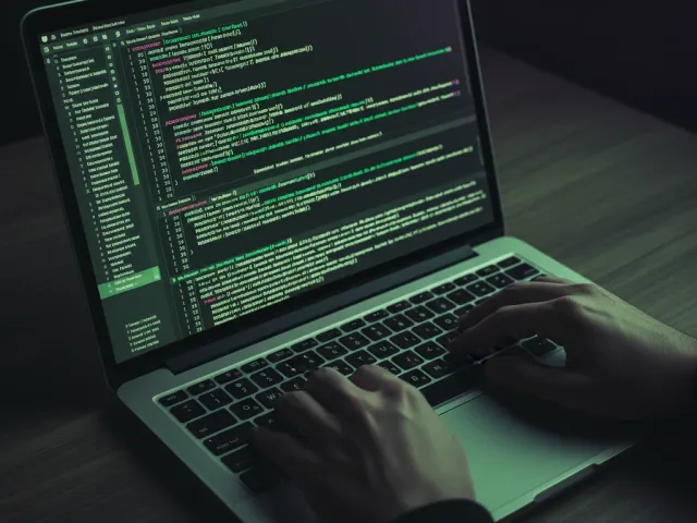 Duas mãos digitando em um teclado de laptop, com código-fonte visível na tela em um ambiente de desenvolvimento. A imagem está com um filtro de tons verdes e roxos escuros, destacando a atividade de programação.