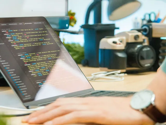 Computador portátil em mesa amadeirada com uma pessoa apoaindo a mão enquanto o computador mostra códigos para programação