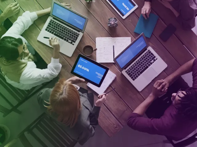 Uma vista aérea de quatro pessoas em uma mesa de madeira, trabalhando em laptops e tablets. Um tablet exibe '85.00%'. A imagem tem um filtro de tons verdes e roxos escuros, transmitindo um ambiente colaborativo e tecnológico de reunião.