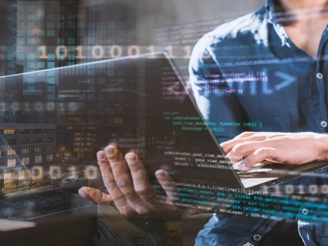 Close-up de um homem vestindo camisa azul segurando um notebook aberto com uma das mãos e digitando com a outra. A imagem apresenta uma sobreposição visual artística com linhas de código de programação (HTML/scripts) e números binários flutuando em azul neon, mesclados com um fundo de edifícios urbanos noturnos iluminados, sugerindo o conceito de desenvolvimento de software e análise de dados no ambiente corporativo.