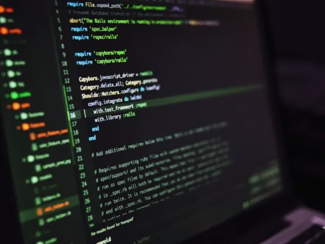 Tela de um laptop exibindo código-fonte em um ambiente de desenvolvimento (possivelmente Ruby on Rails), com linhas de código em tons verdes e roxos escuros, ilustrando a parte técnica do desenvolvimento de software.
